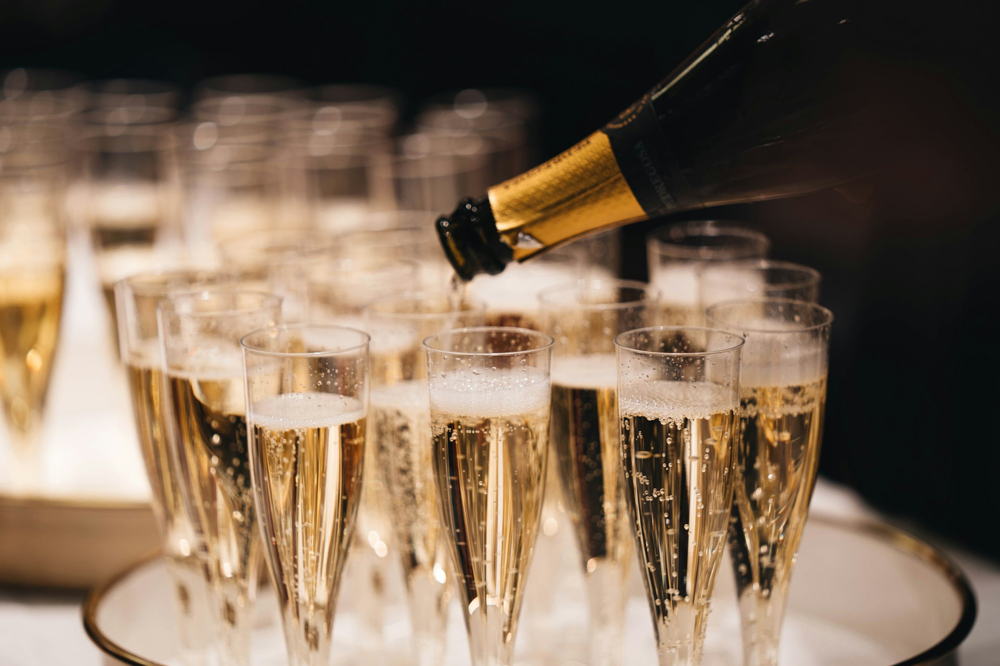 Champagne being poured into a tray of filled flutes against a dark background