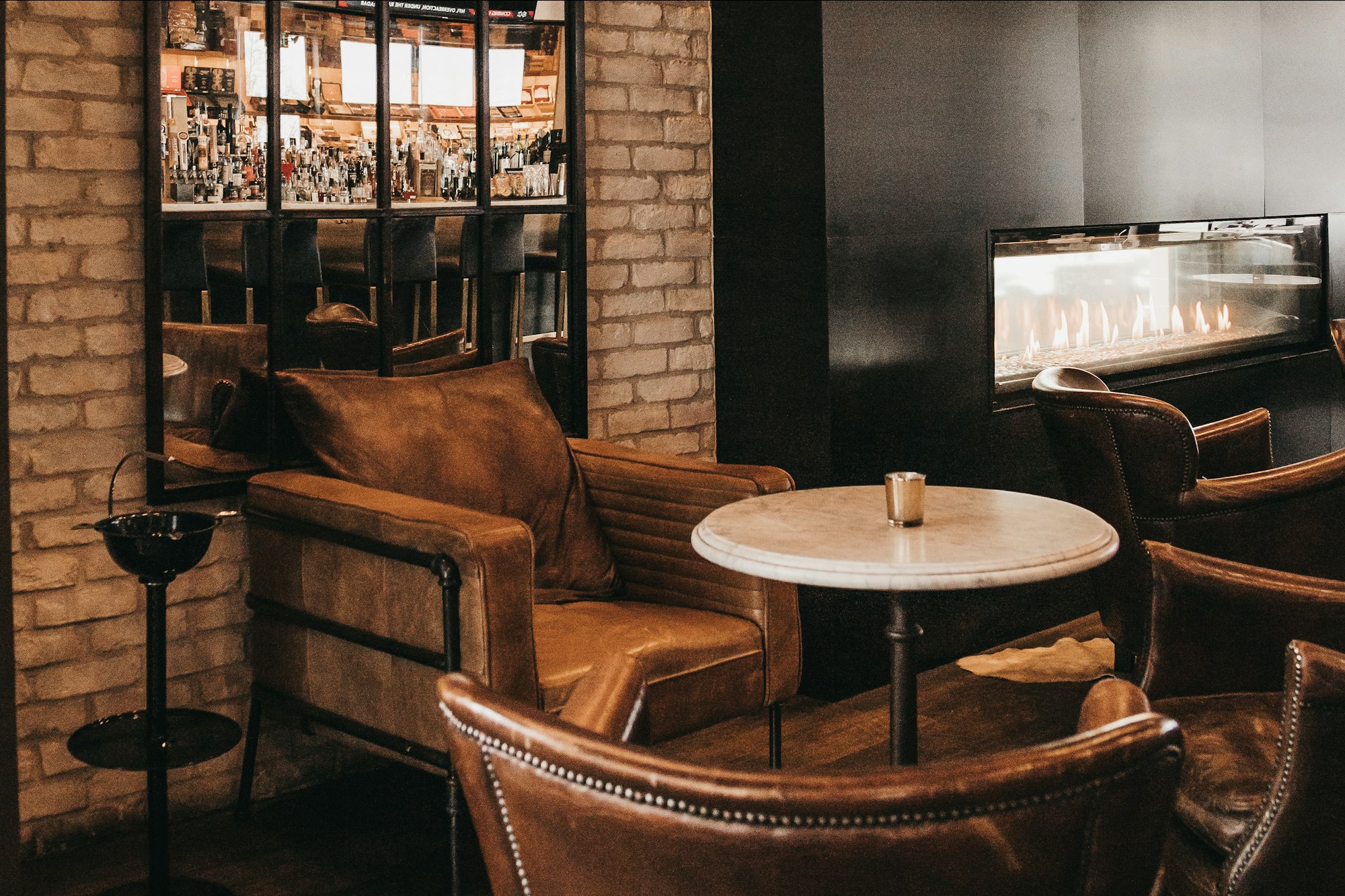 A dark lounge of worn leather armchairs around a marble table, a long gas fire burning in the wall behind them