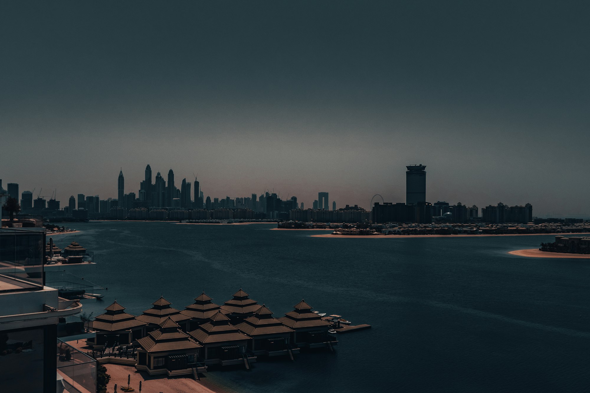 A Gulf shoreline at dusk, a city skyline silhouetted on the horizon under an amber band of light above dark water