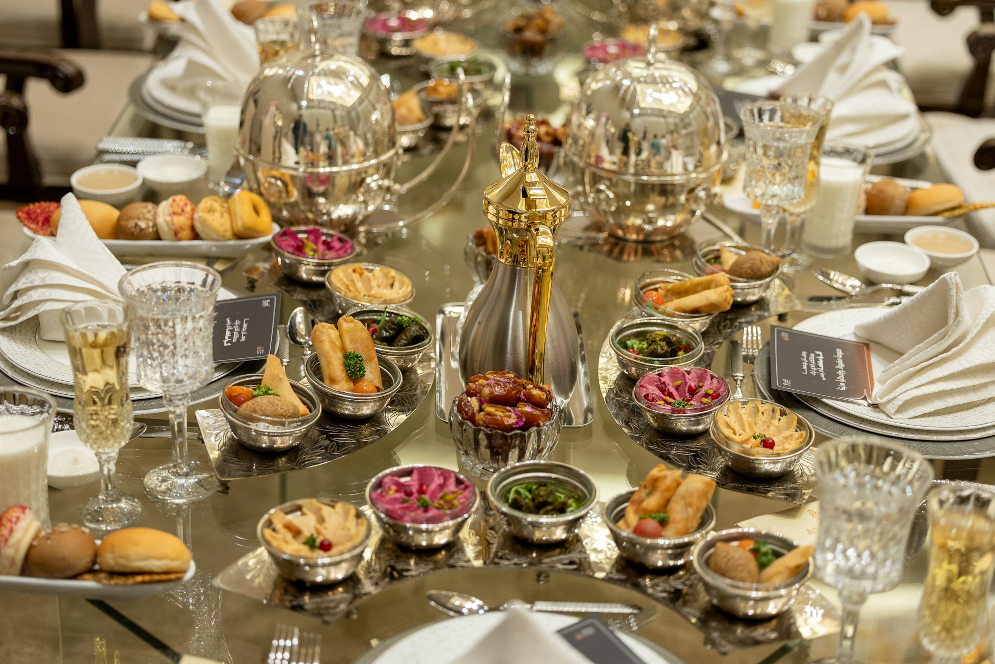 A long table laid for iftar with silver domed dishes, a gold dallah coffee pot, dates and mezze in silver bowls
